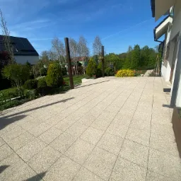 Jasna, kwadratowa kostka brukowa tworząca taras przylegający do domu z ogrodem, widoczne drewniane słupki pergoli, roślinność i błękitne niebo.