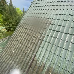 ABC Mycie Wrocław - Zielony dach z blachy falistej, częściowo pokryty mchem i zabrudzeniami, czyszczony strumieniem wody pod ciśnieniem, z widocznym fragmentem lancy myjki ciśnieniowej w dolnej części kadru, na tle...