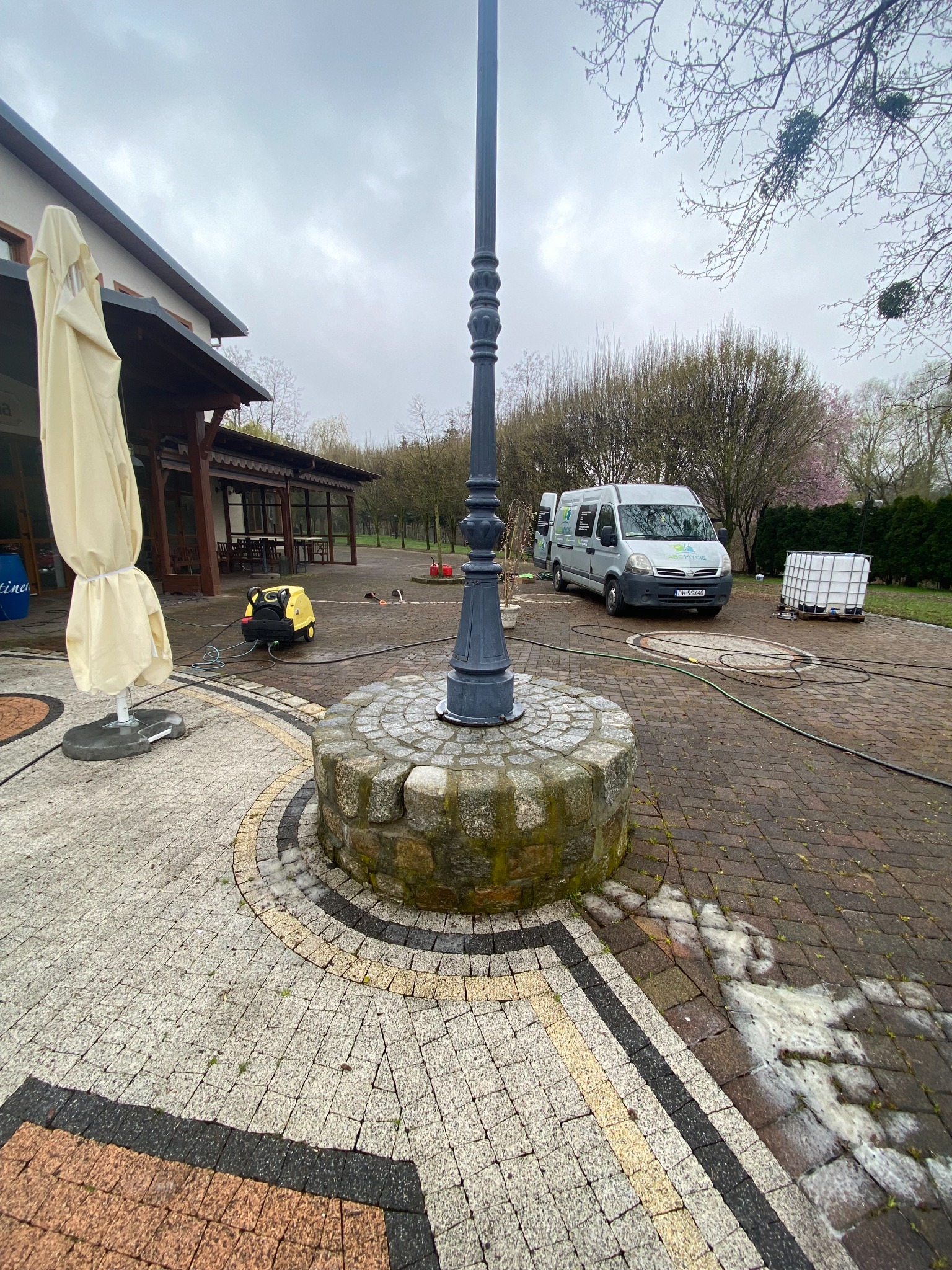 Mycie kostki brukowej na zewnątrz budynku z widocznym słupem oświetleniowym, samochodem dostawczym z napisem 'ABC Mycie' oraz generatorem ciśnieniowym.