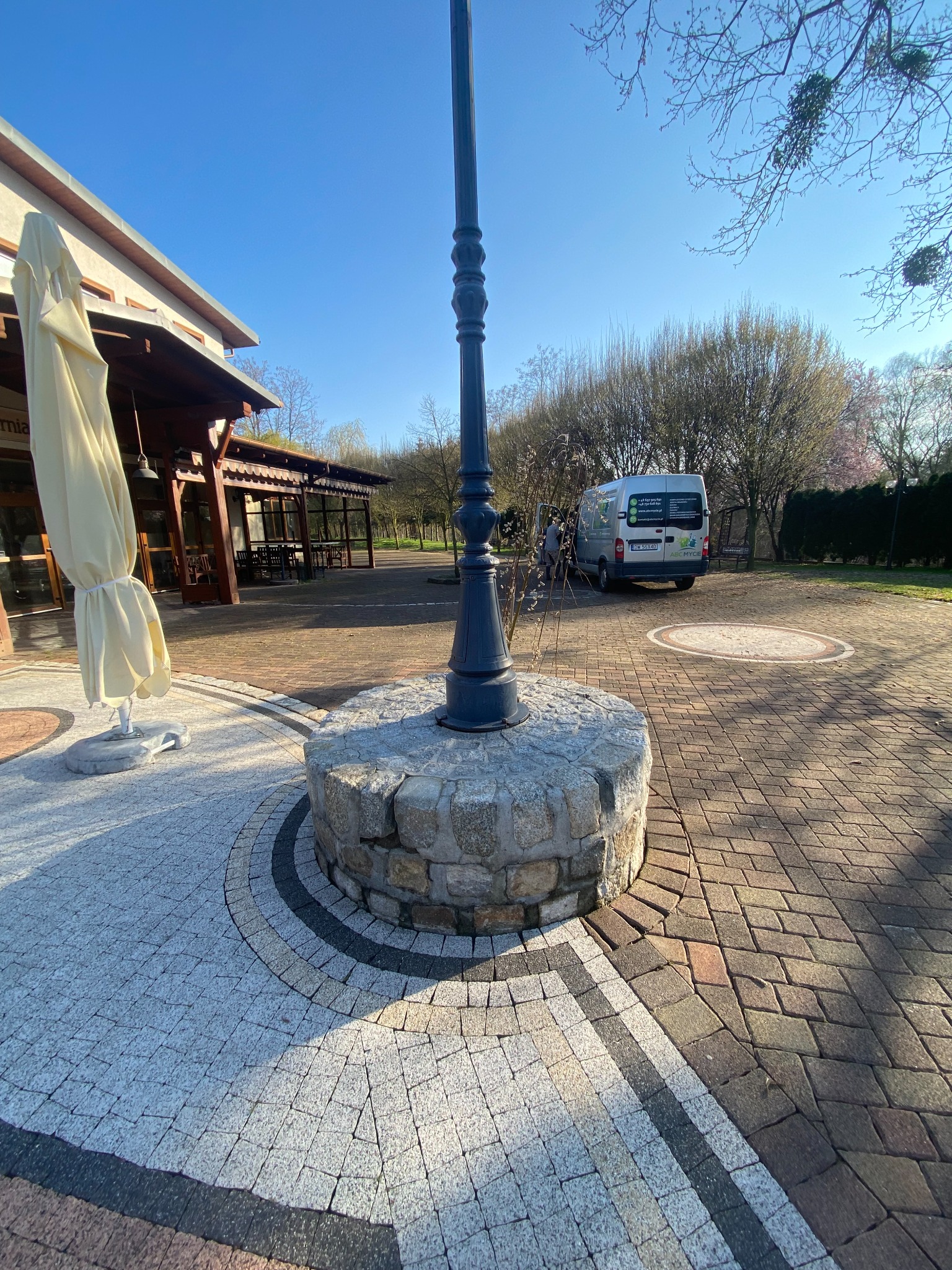 Ułożona z kostki brukowej powierzchnia placu z ozdobnymi wzorami, w tle budynek z zadaszeniem i zaparkowany bus z widocznym logo firmy.