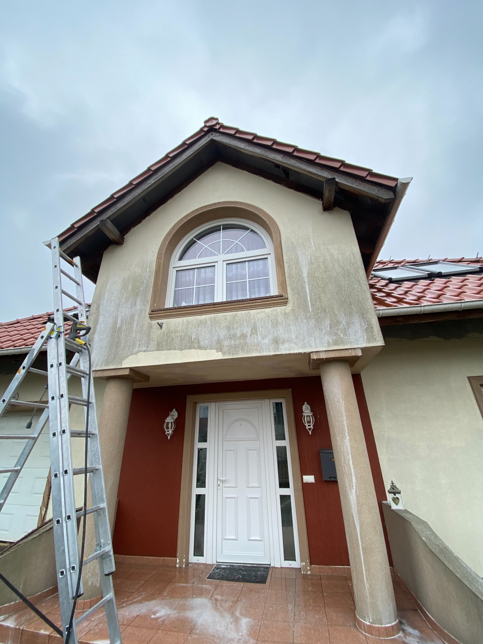 Dom z częściowo umytą elewacją, widoczne zacieki i różnica w kolorze, drabina oparta o ścianę, pochmurne niebo.