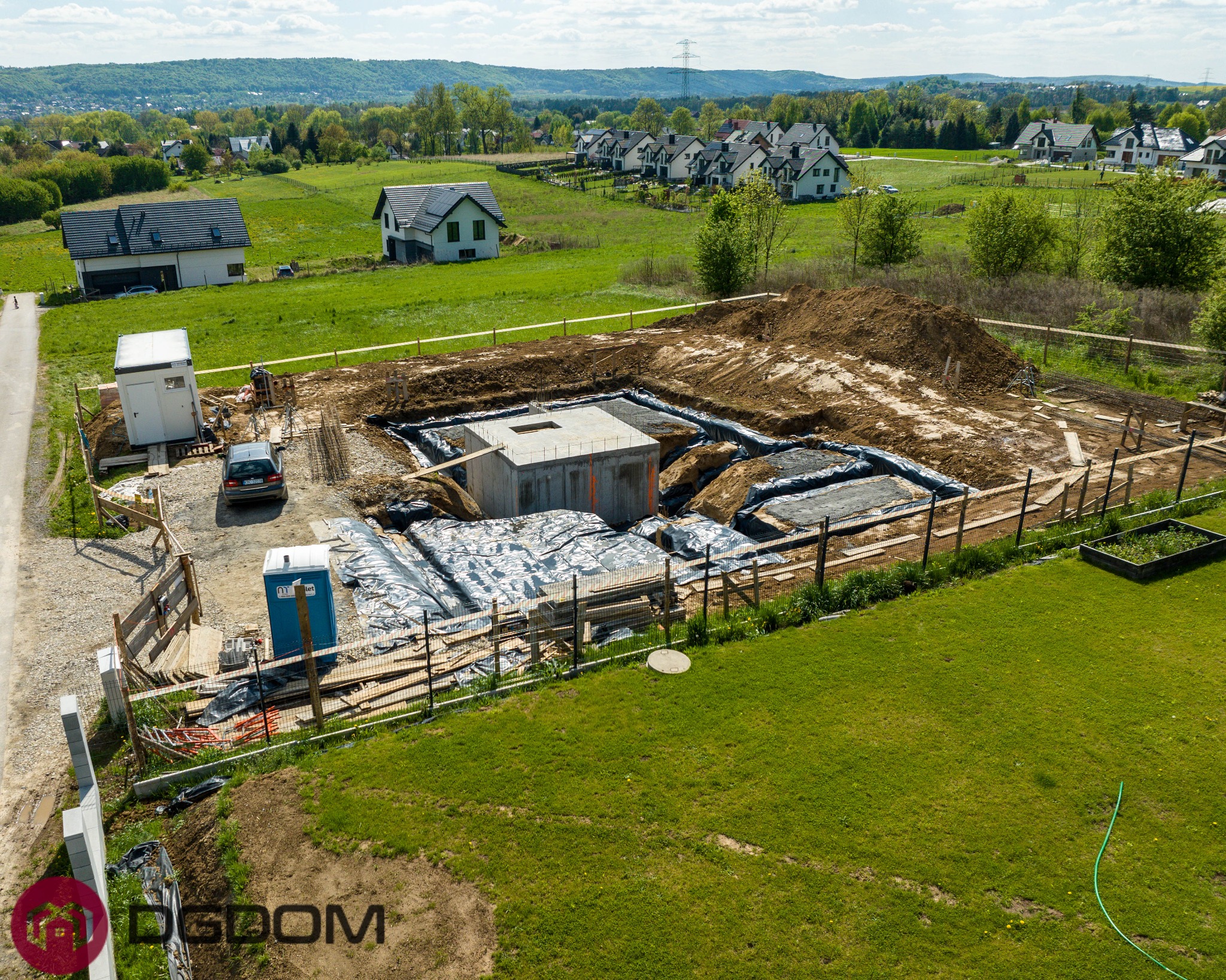 Fundamenty domu w trakcie budowy, zabezpieczone folią, z widocznym szalunkiem i zbrojeniem. W tle zabudowa jednorodzinna i zieleń.