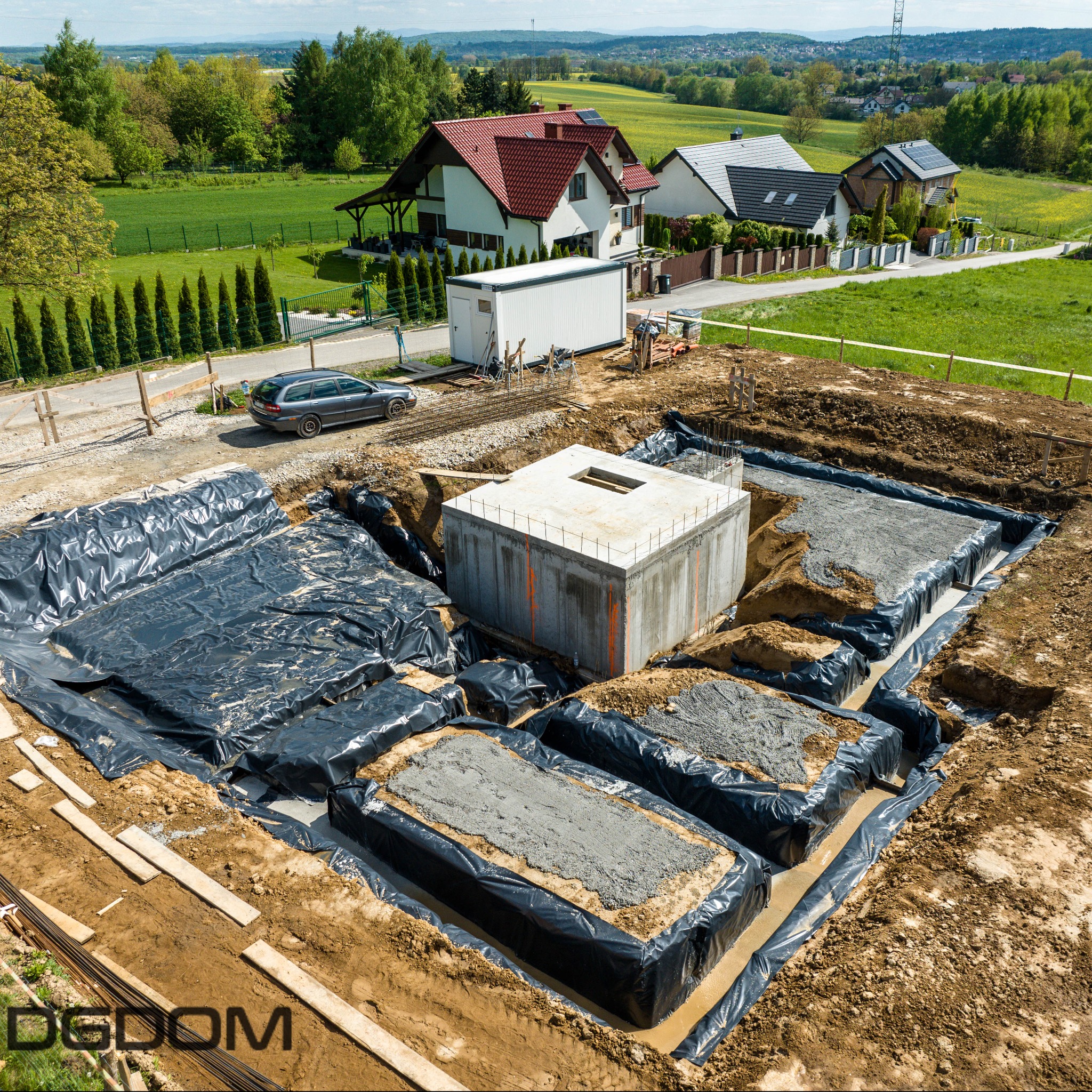 Fundamenty domu w trakcie budowy, zabezpieczone folią, z widocznym zbrojeniem i betonową studzienką. W tle krajobraz z domami i zielenią.