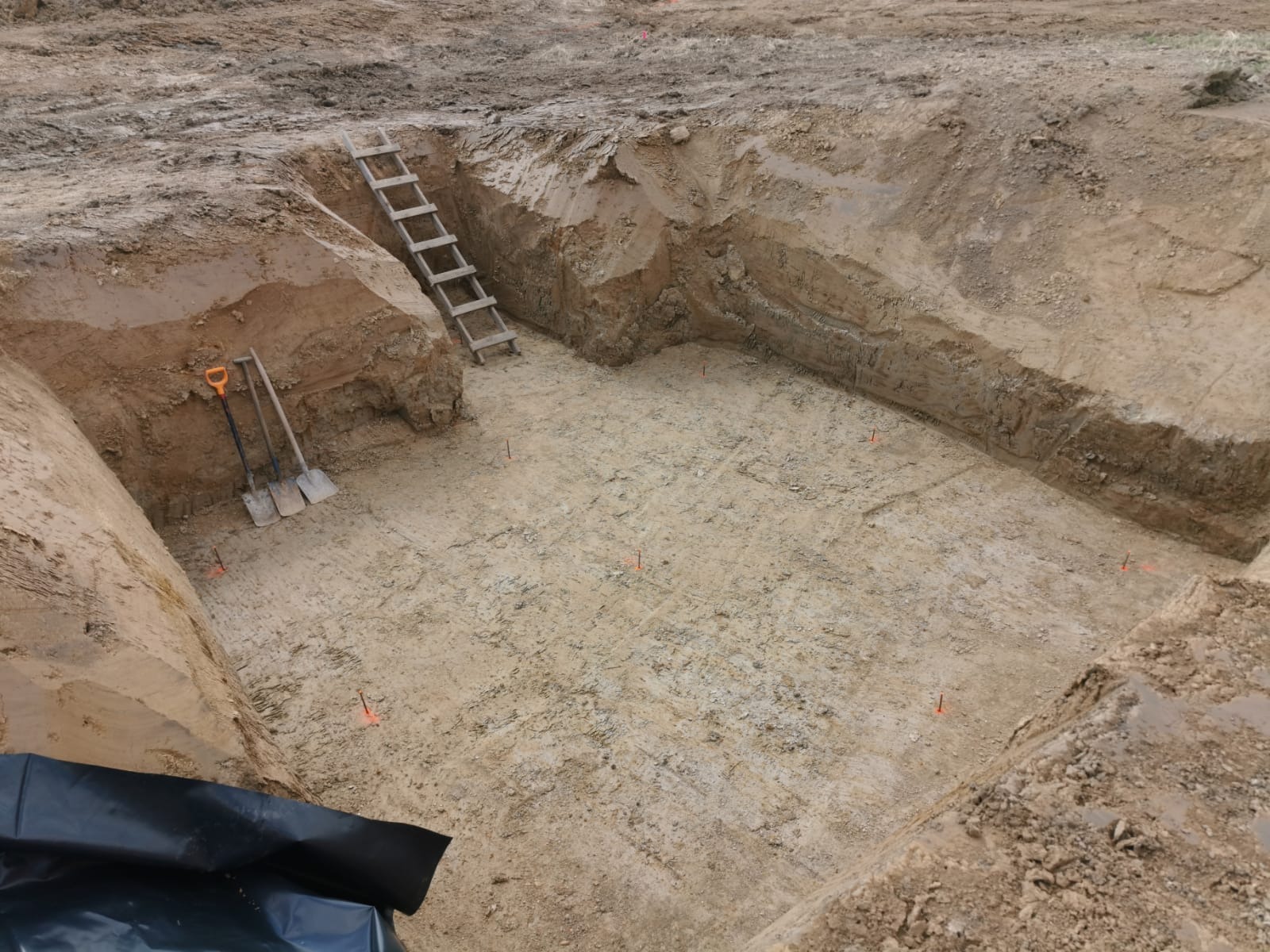 Wykop pod fundamenty domu z widocznymi śladami po szpadlu, drewnianą drabiną i pomarańczowymi znacznikami. Na pierwszym planie czarna folia budowlana.