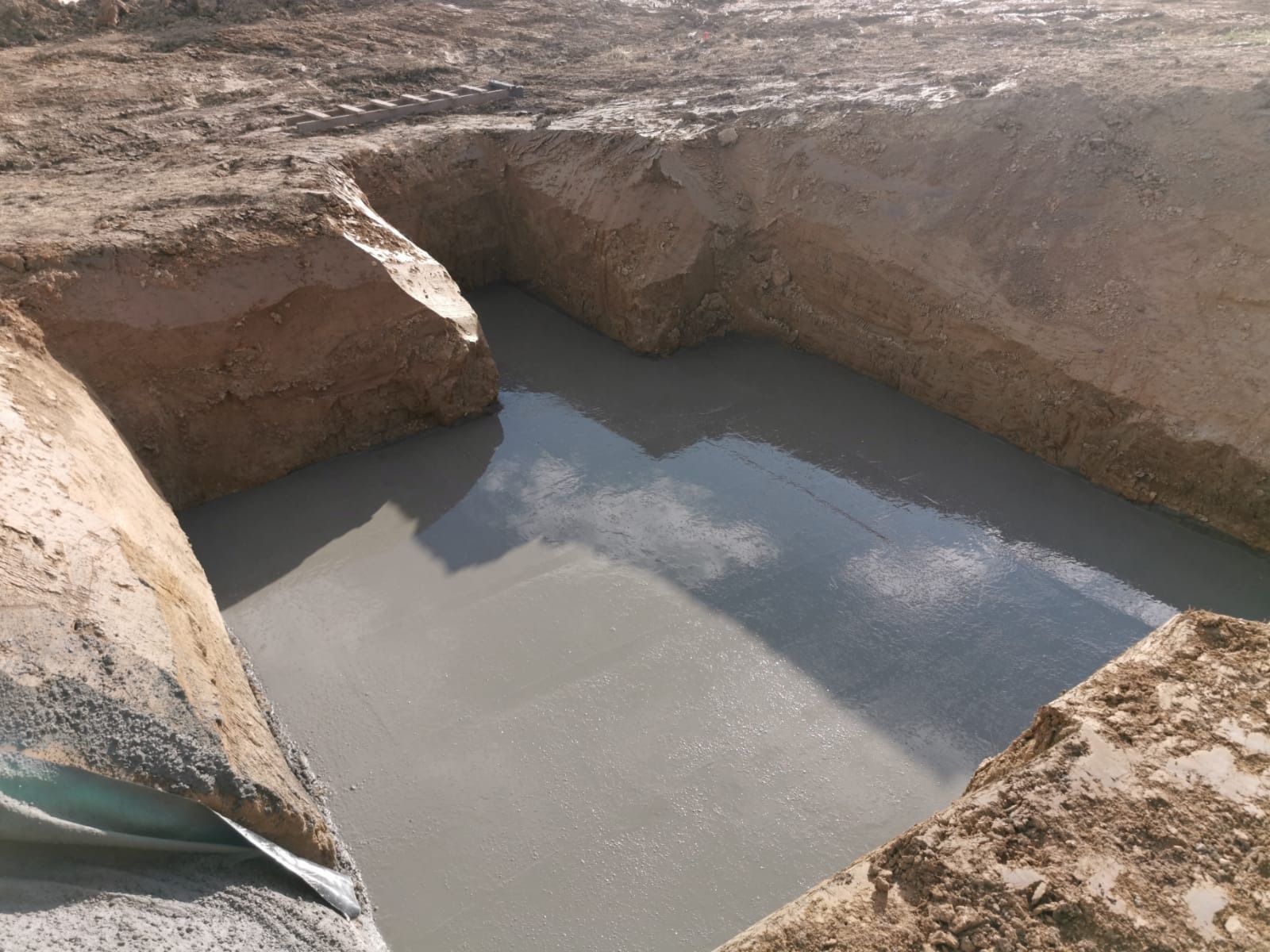 Zalane fundamenty pod dom murowany, widoczne ściany wykopu z ziemi i odbicie nieba w mokrym betonie. W tle drabina.