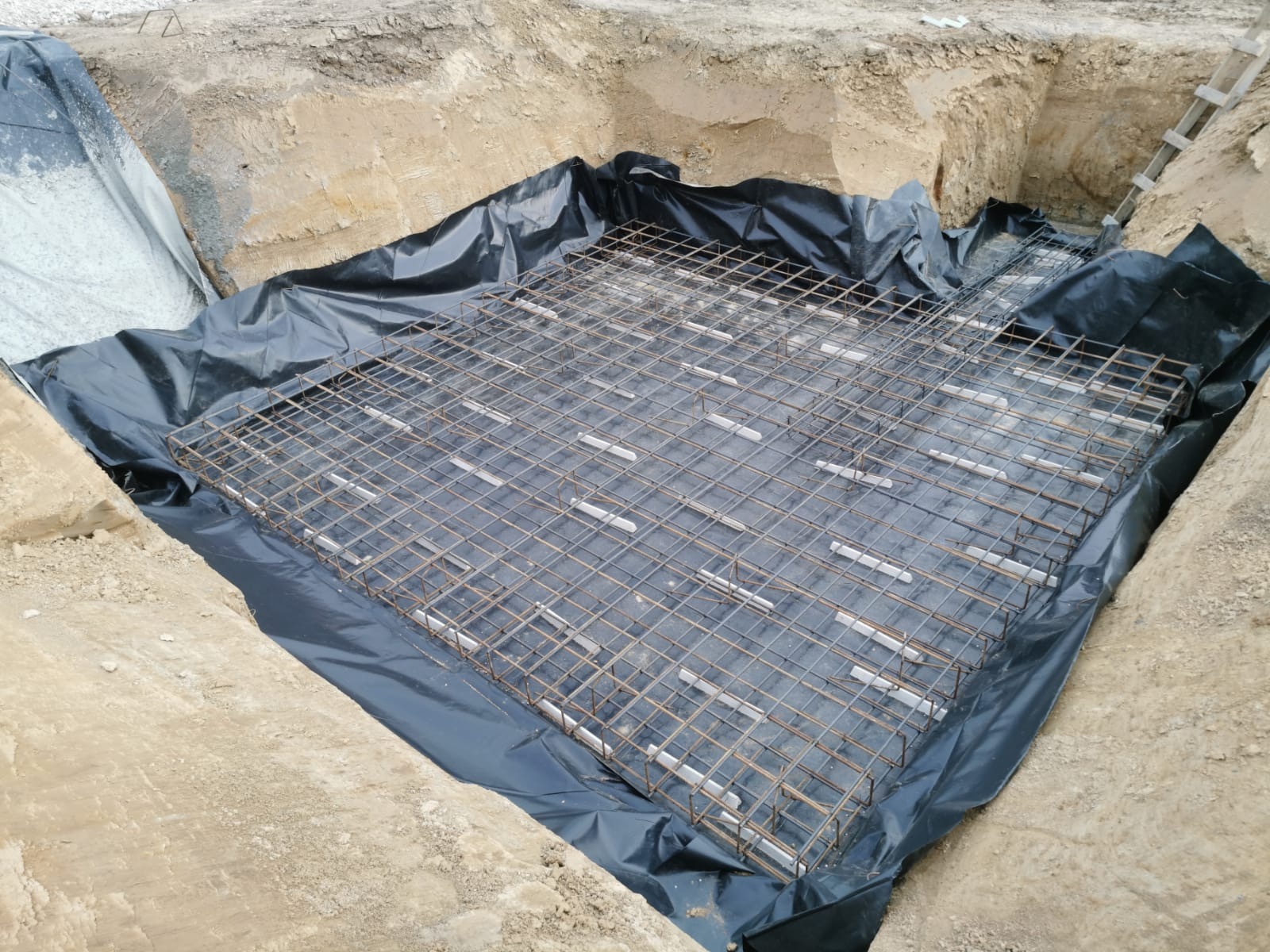 Zbrojenie fundamentu domu jednorodzinnego, ułożone na czarnej folii w wykopie. Widoczna drabina oparta o ścianę wykopu. Etap budowy domu.