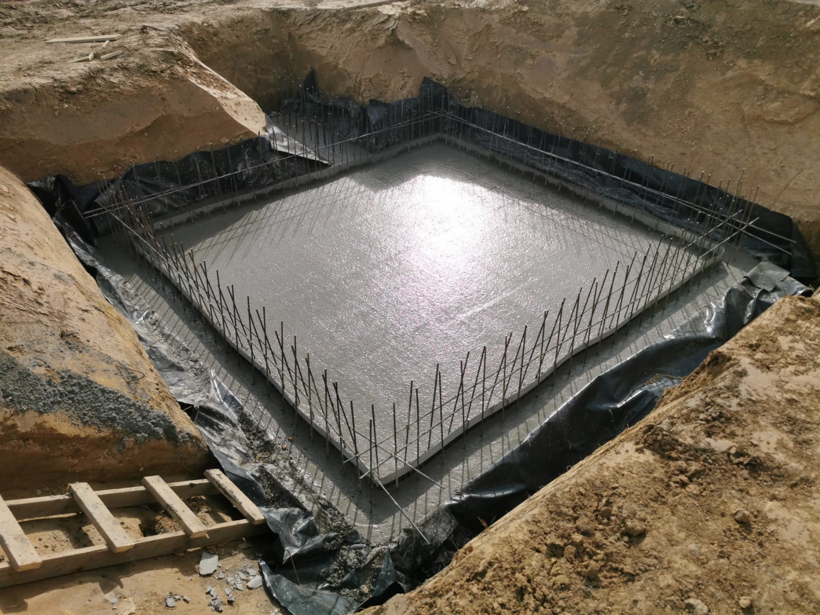 Zalany fundament pod dom z wystającymi prętami zbrojeniowymi, zabezpieczony czarną folią, widok z góry na wykop z ziemią i drewnianą drabinką.