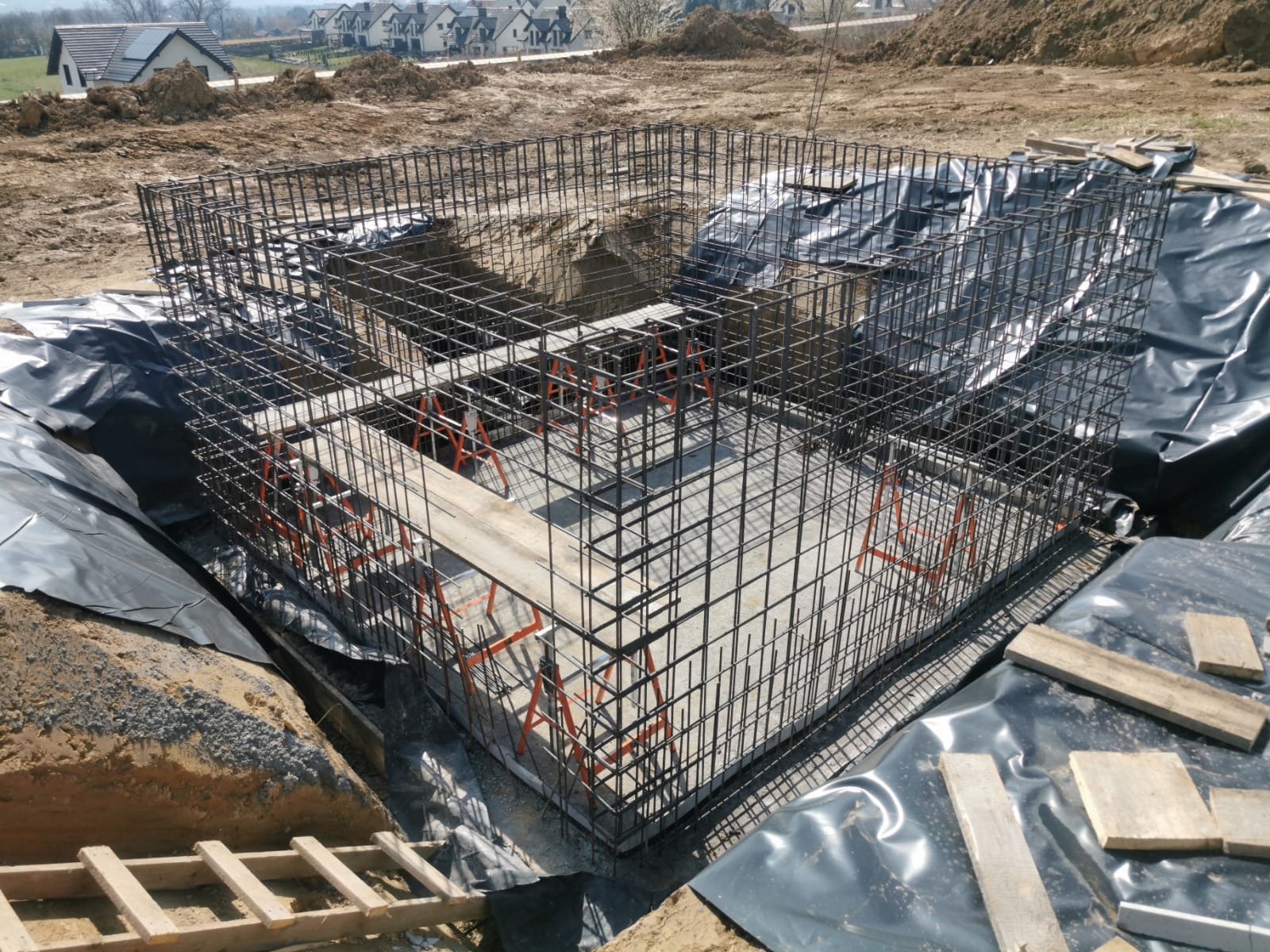 Zbrojenie fundamentów domu jednorodzinnego, widoczne pomarańczowe podpory i czarna folia izolacyjna. W tle zabudowa jednorodzinna i ziemia.