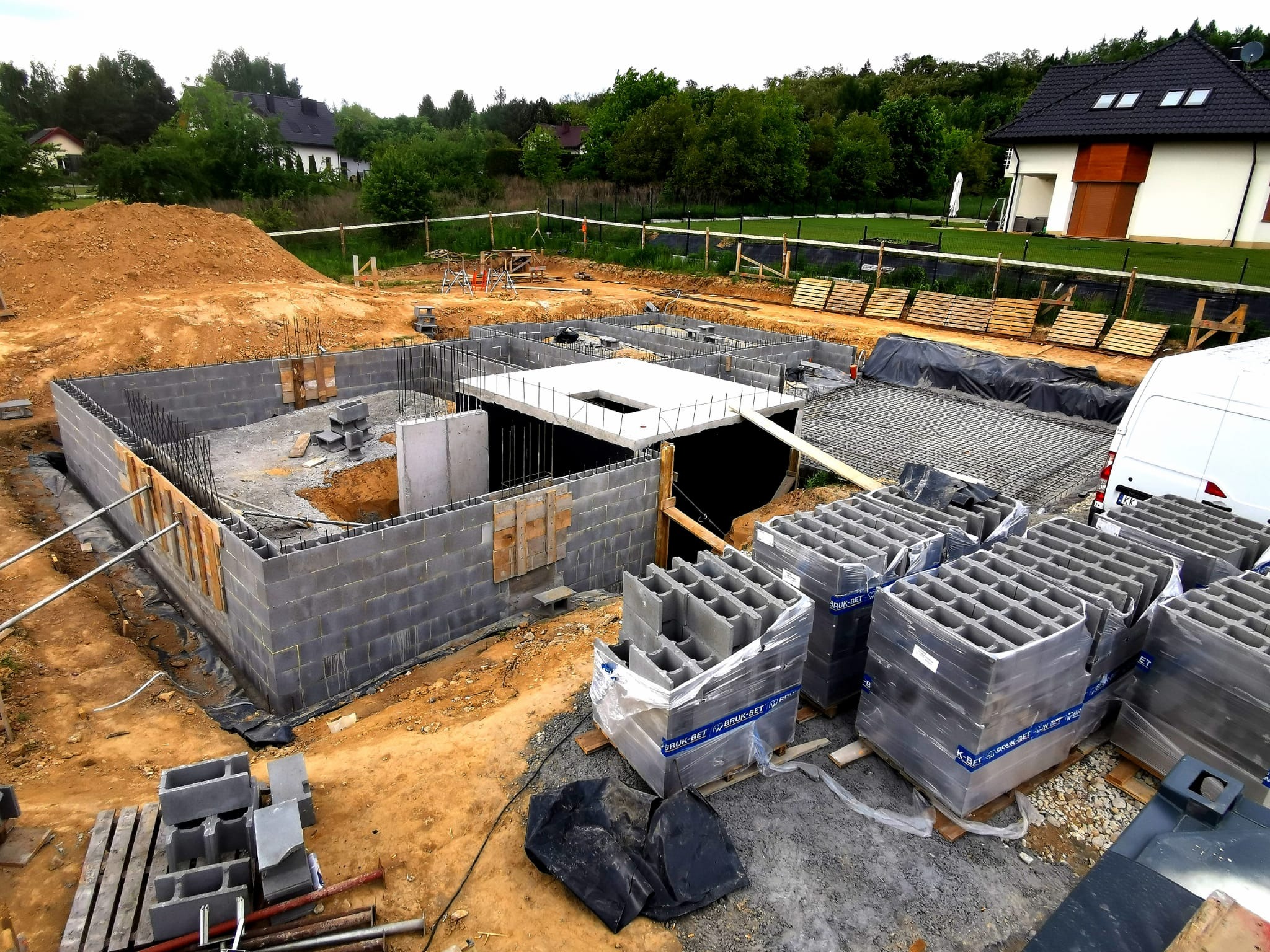 Budowa domu: fundamenty z bloczków betonowych, zbrojenie, przygotowany teren. Widok z góry na plac budowy w otoczeniu zieleni i zabudowy jednorodzinnej.