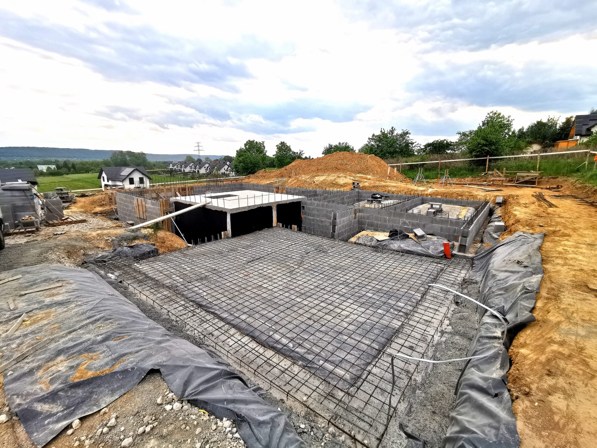 Budowa domu: zbrojenie fundamentów z widocznymi bloczkami betonowymi, folią ochronną i ziemią. W tle zabudowa jednorodzinna i pochmurne niebo.