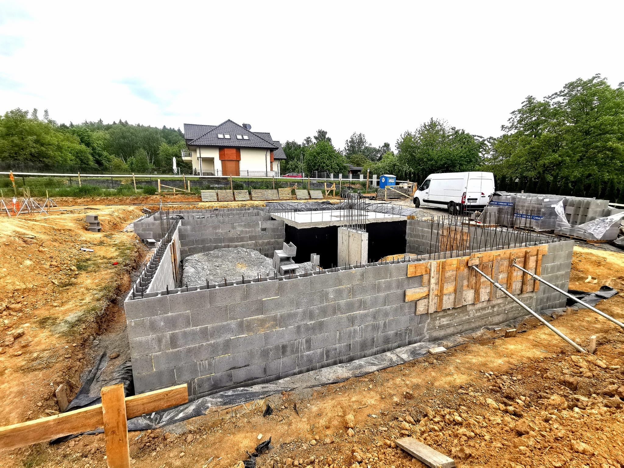 Fundamenty domu z bloczków betonowych, widoczne zbrojenia i szalunki. W tle dom jednorodzinny i biały bus dostawczy. Etap budowy domu.