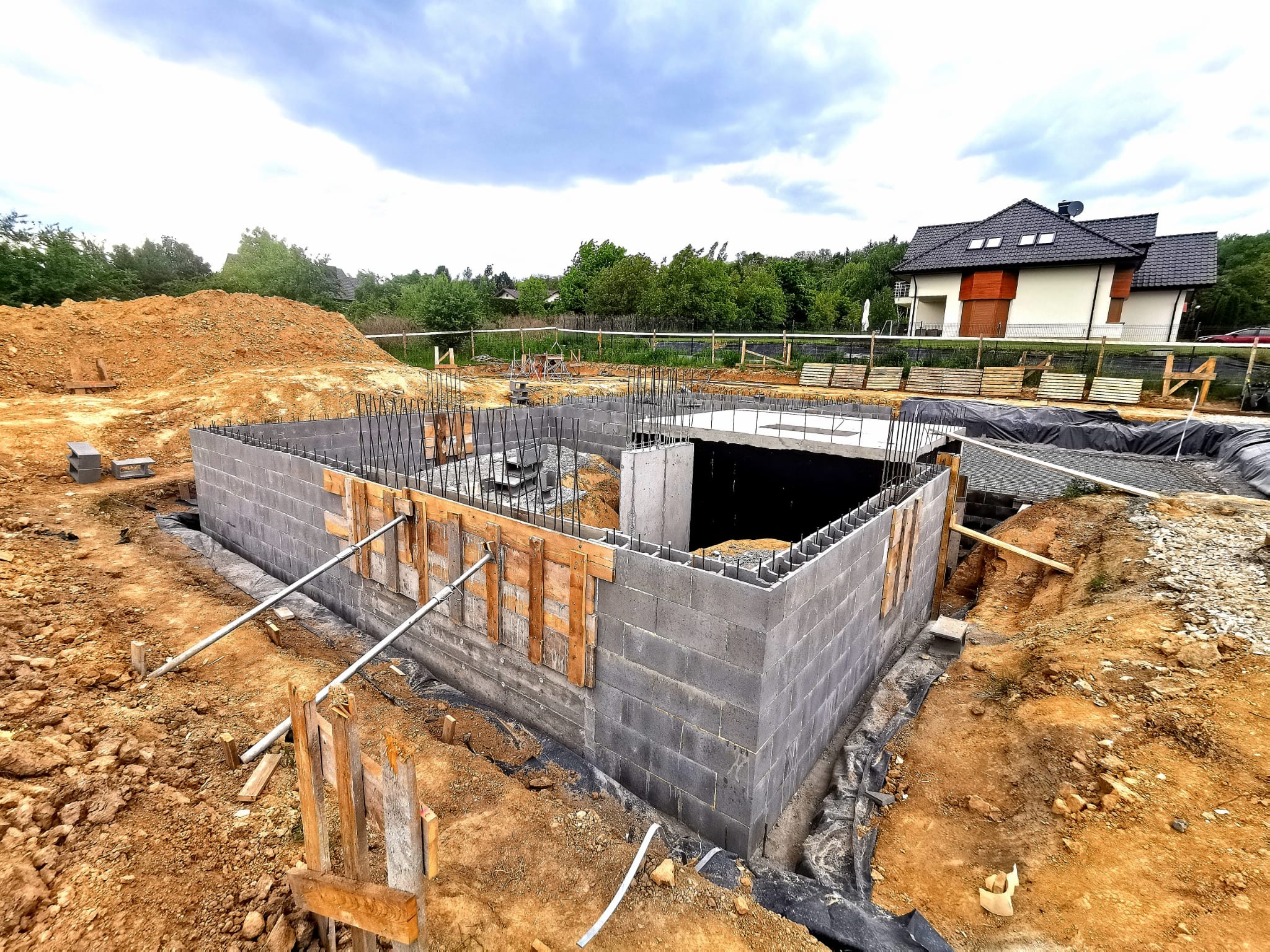 Fundamenty domu z betonu komórkowego, zbrojenie pionowe, szalunki drewniane, widok na budynek w tle, teren budowy w trakcie prac ziemnych.