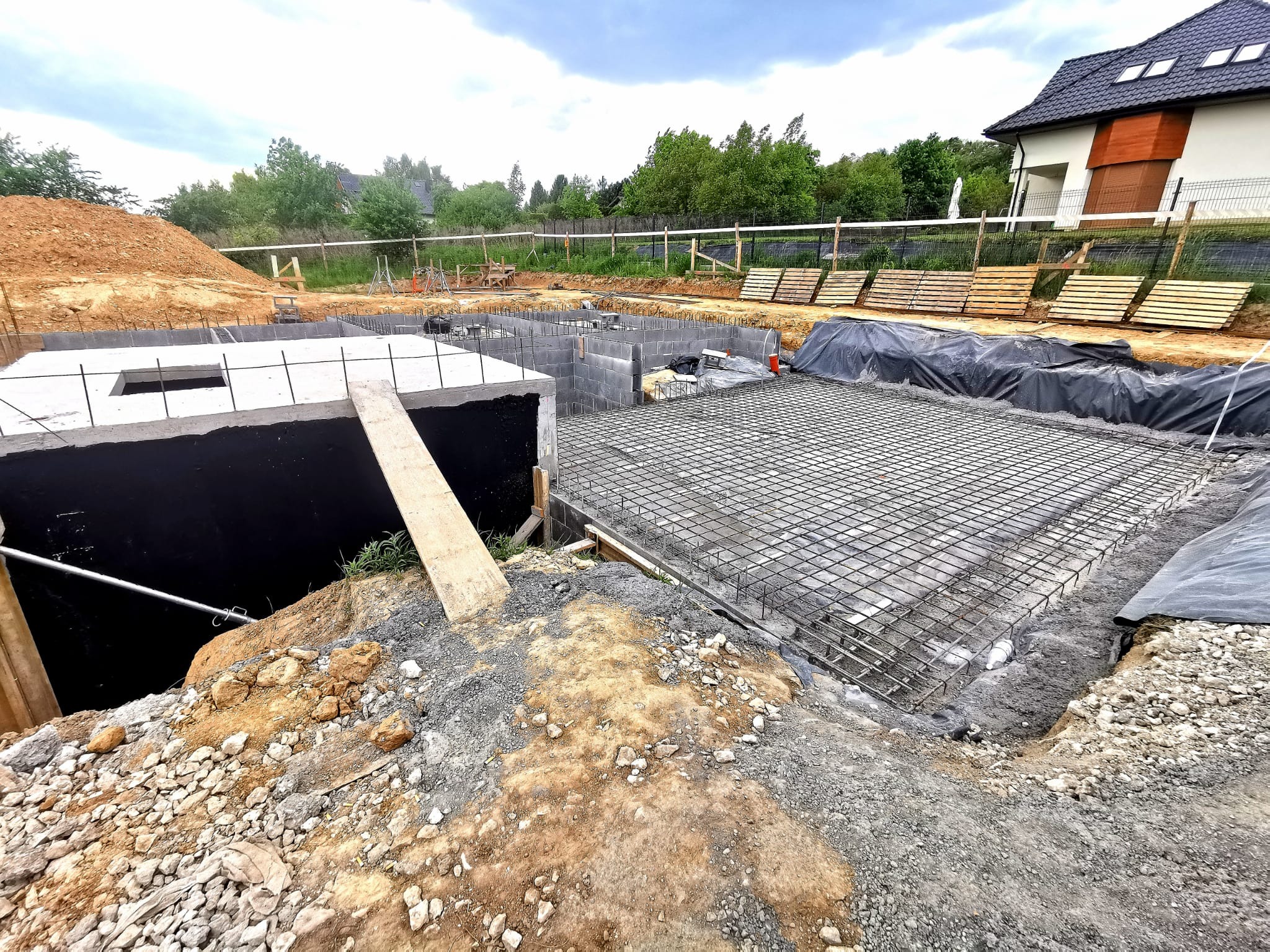 Fundamenty domu w trakcie budowy: zbrojenie, betonowe ściany, wykop ziemny. Widoczne palety i fragment domu w tle. Ujęcie z góry, szeroki kadr.