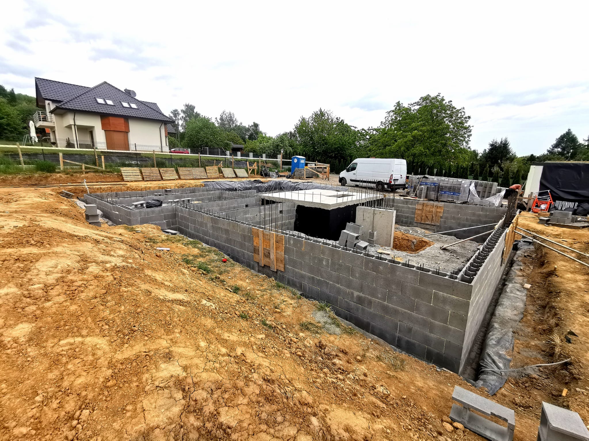 Fundamenty domu z bloczków betonowych na działce, widoczny zarys budynku i zbrojenia. W tle dom jednorodzinny i samochód dostawczy.