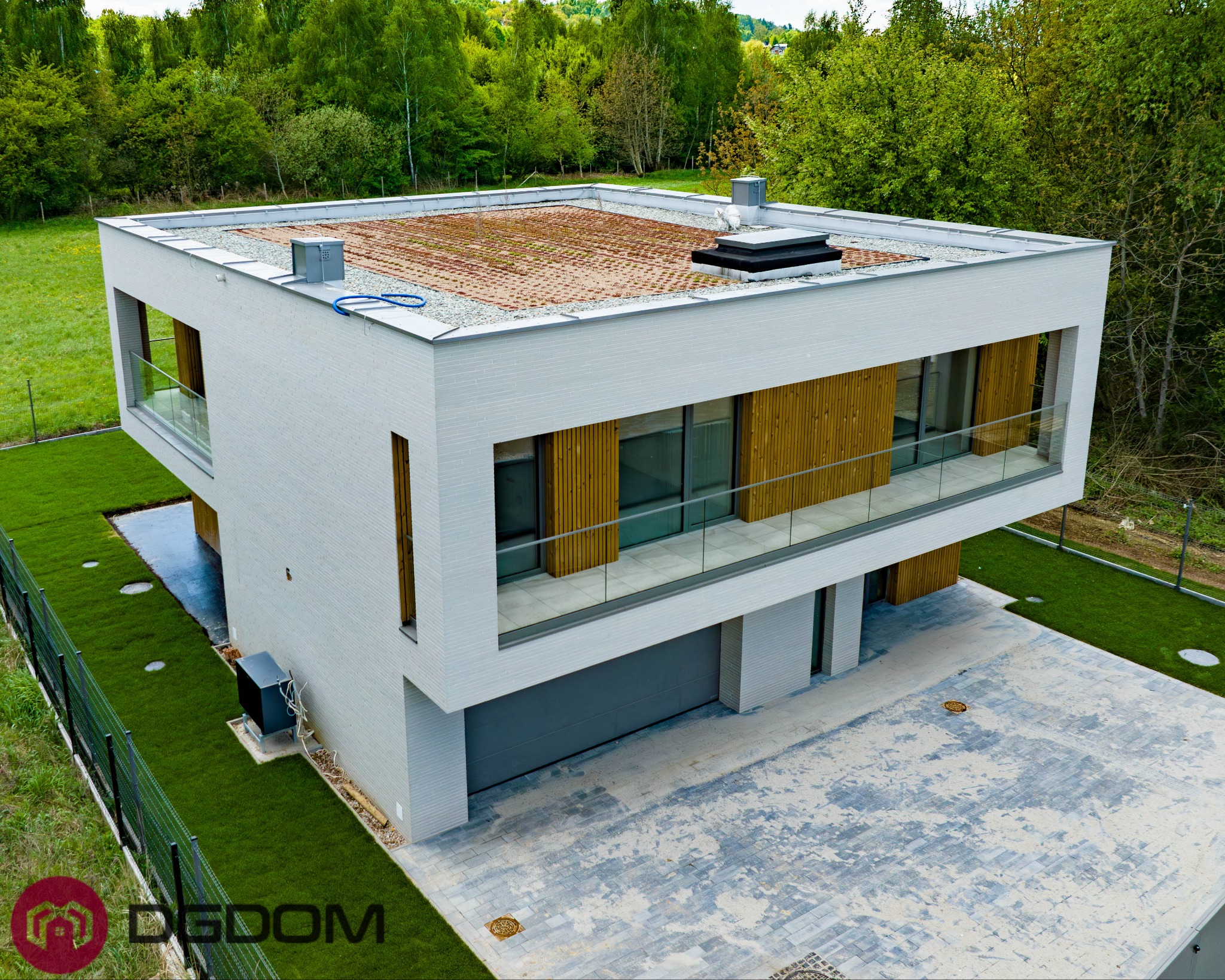 Nowoczesny dom murowany z płaskim dachem porośniętym roślinnością, widok z góry. Biała elewacja, drewniane elementy, szklane balustrady. Zadbany trawnik i kostka brukowa.