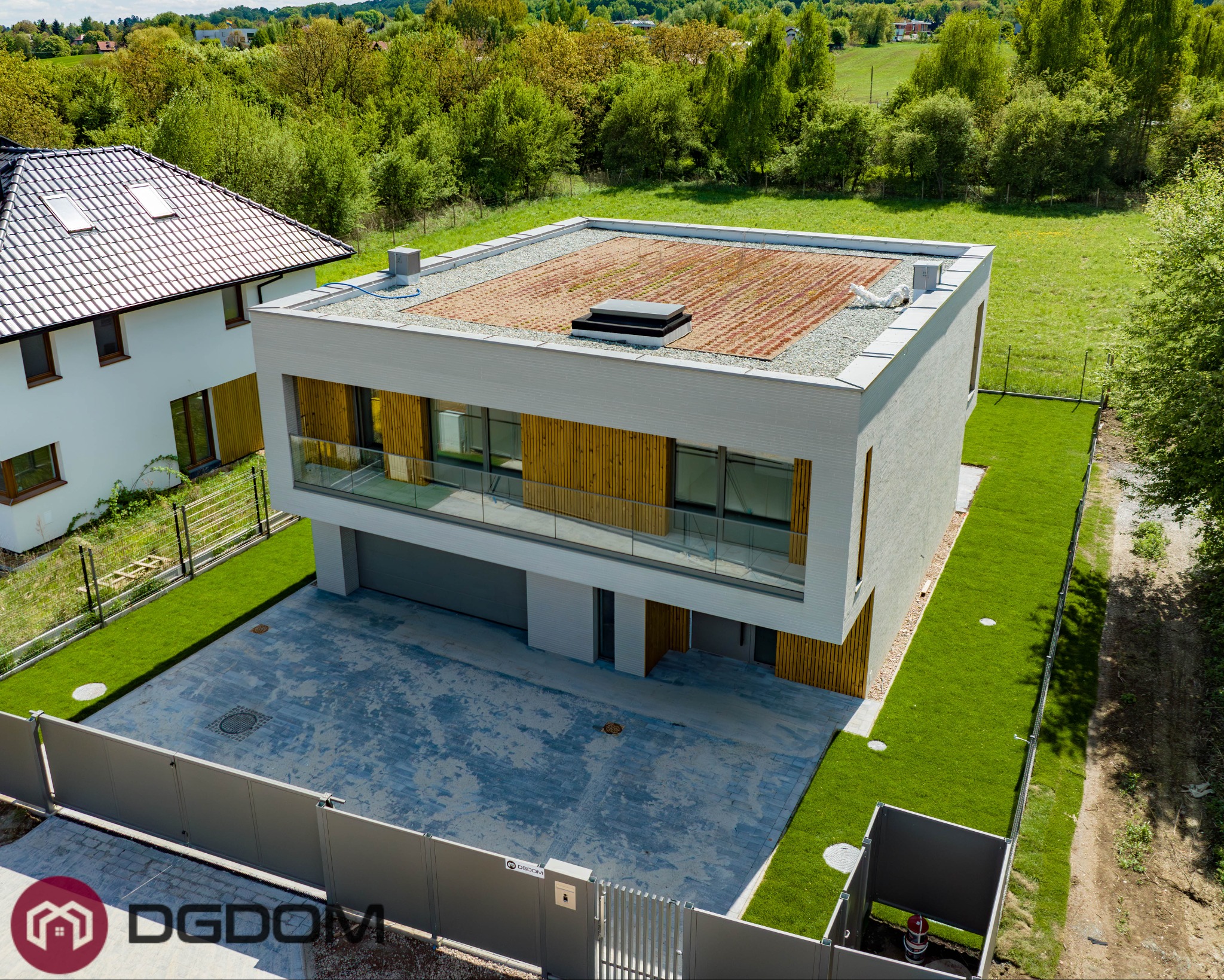 Nowoczesny dom murowany z zielonym dachem, widok z góry. Minimalistyczna bryła, drewniane akcenty elewacji, zadbany trawnik i szara kostka brukowa na podjeździe.