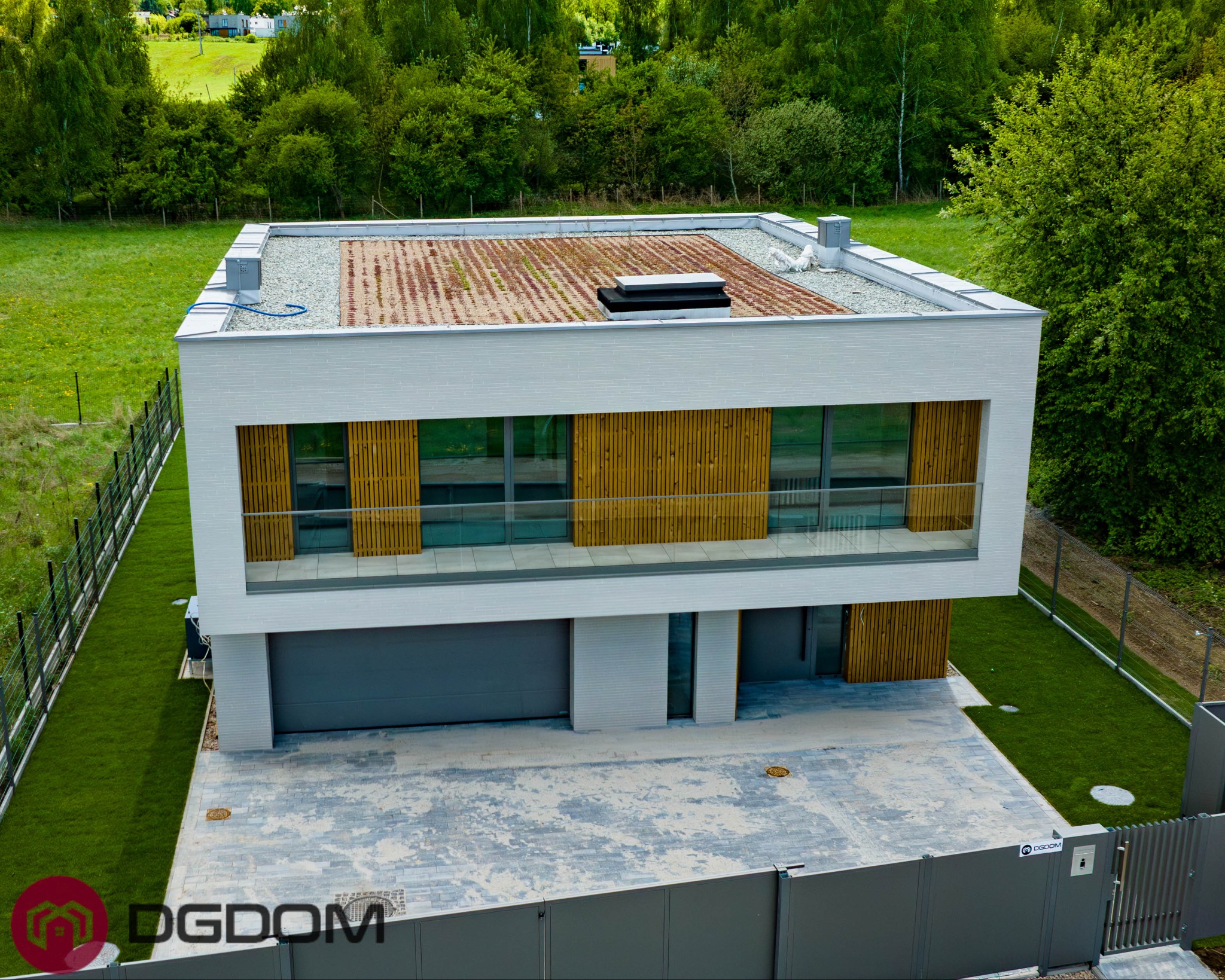 Nowoczesny dom z płaskim dachem pokrytym roślinnością, białą elewacją i drewnianymi elementami, widziany z góry, otoczony zielenią i ogrodzeniem.