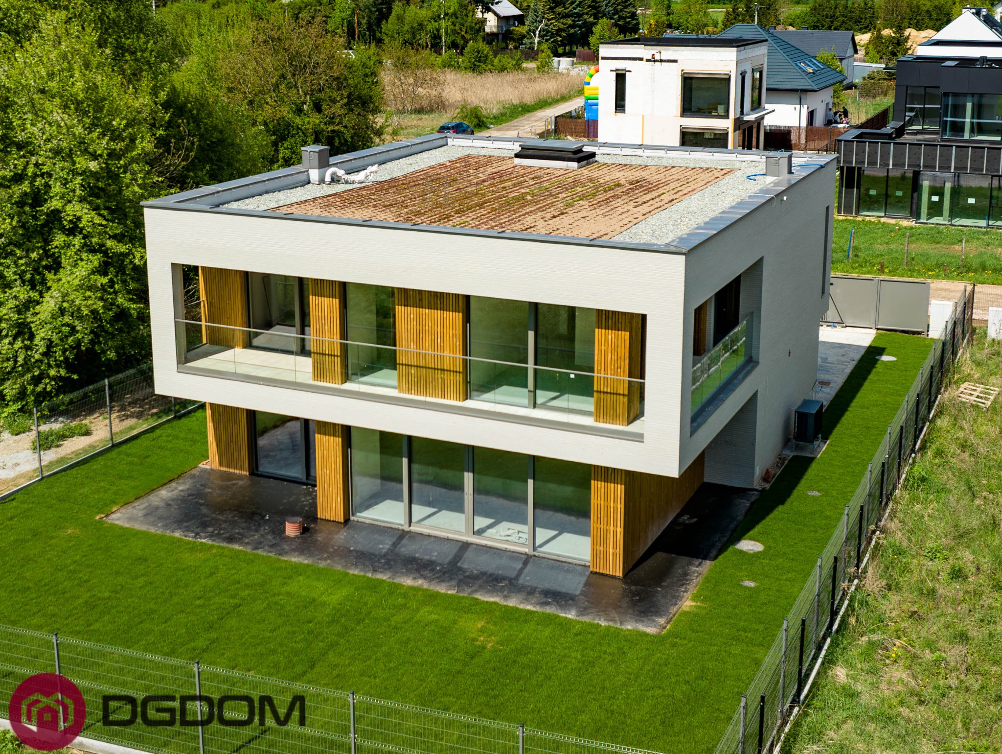 Nowoczesny dom murowany z płaskim dachem porośniętym roślinnością, widok z góry, ogrodzony siatką. Minimalistyczna architektura z drewnianymi elementami elewacji.