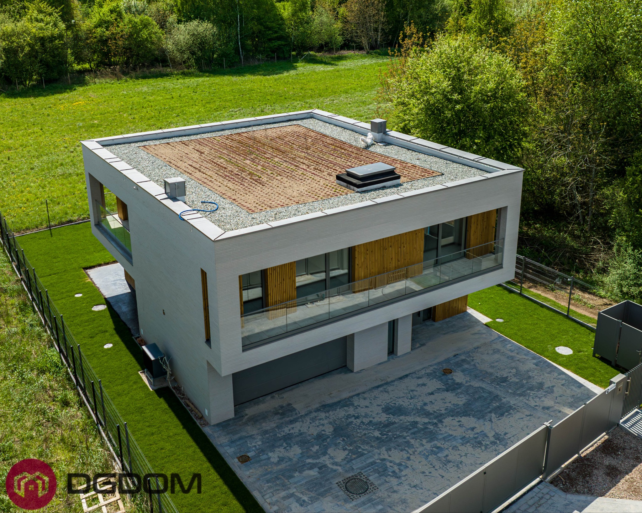 Nowoczesny dom murowany z zielonym dachem, widok z góry. Minimalistyczna bryła, drewniane akcenty, szara kostka brukowa i zadbany trawnik. Ogrodzenie panelowe.