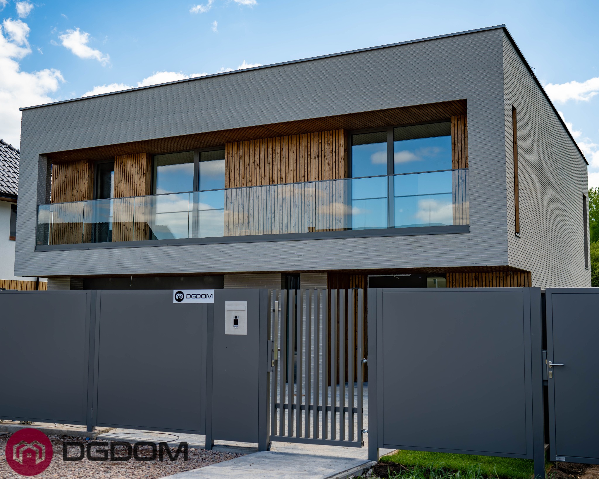 Dwukondygnacyjny dom murowany z szarej cegły z drewnianymi elementami wykończeniowymi i szklaną balustradą balkonu, widziany zza grafitowego ogrodzenia.