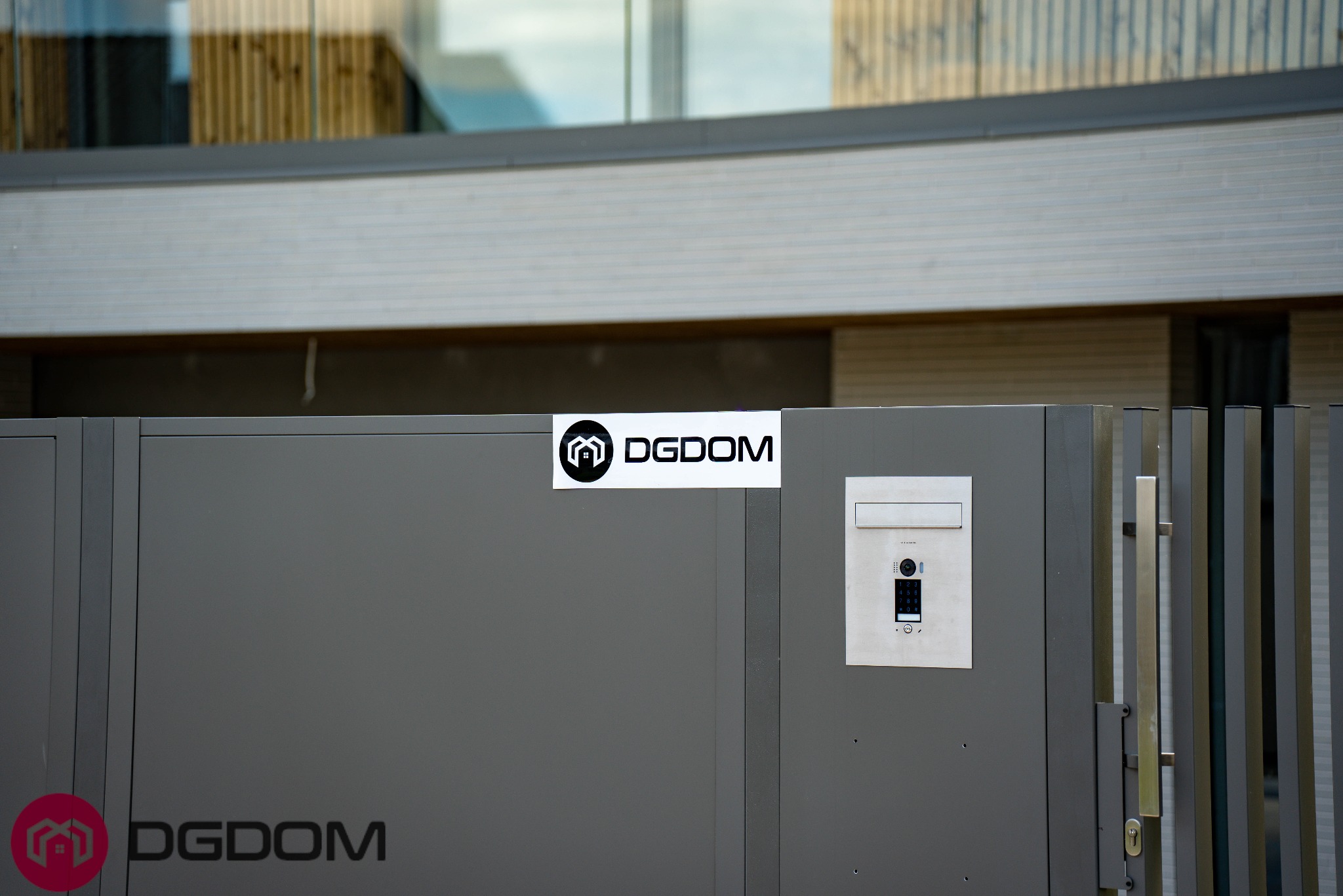 Szara brama z domofonem i naklejką z logo firmy budowlanej DGDOM, na tle fragmentu elewacji domu z drewnianymi elementami. Nowoczesny design.
