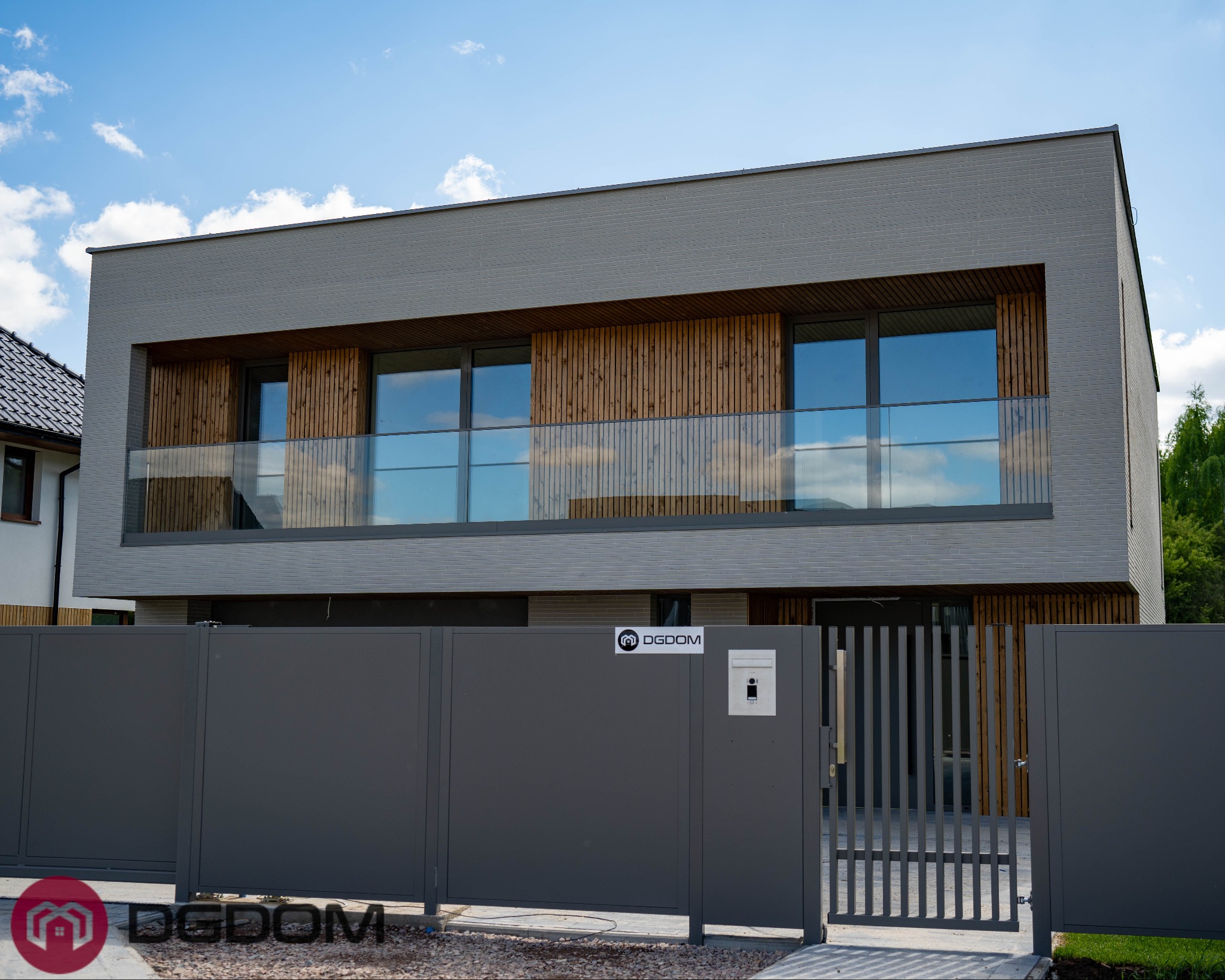 Nowoczesny dom murowany z szarą elewacją i drewnianymi akcentami, widoczny zza grafitowego ogrodzenia z logo firmy budowlanej. Błękitne niebo w odbiciu szyb.