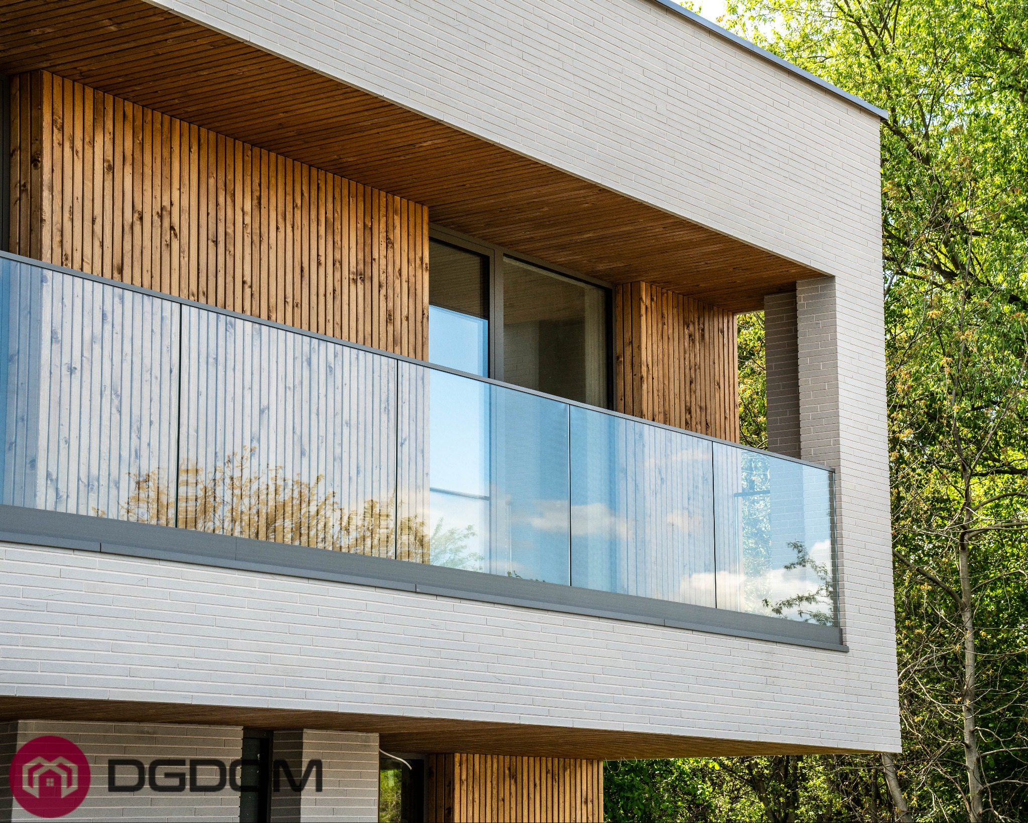 Nowoczesny dom murowany z balkonem o szklanej balustradzie, wykończony drewnem i jasną cegłą, otoczony zielenią. Widok z dołu, podkreślający architekturę budynku.