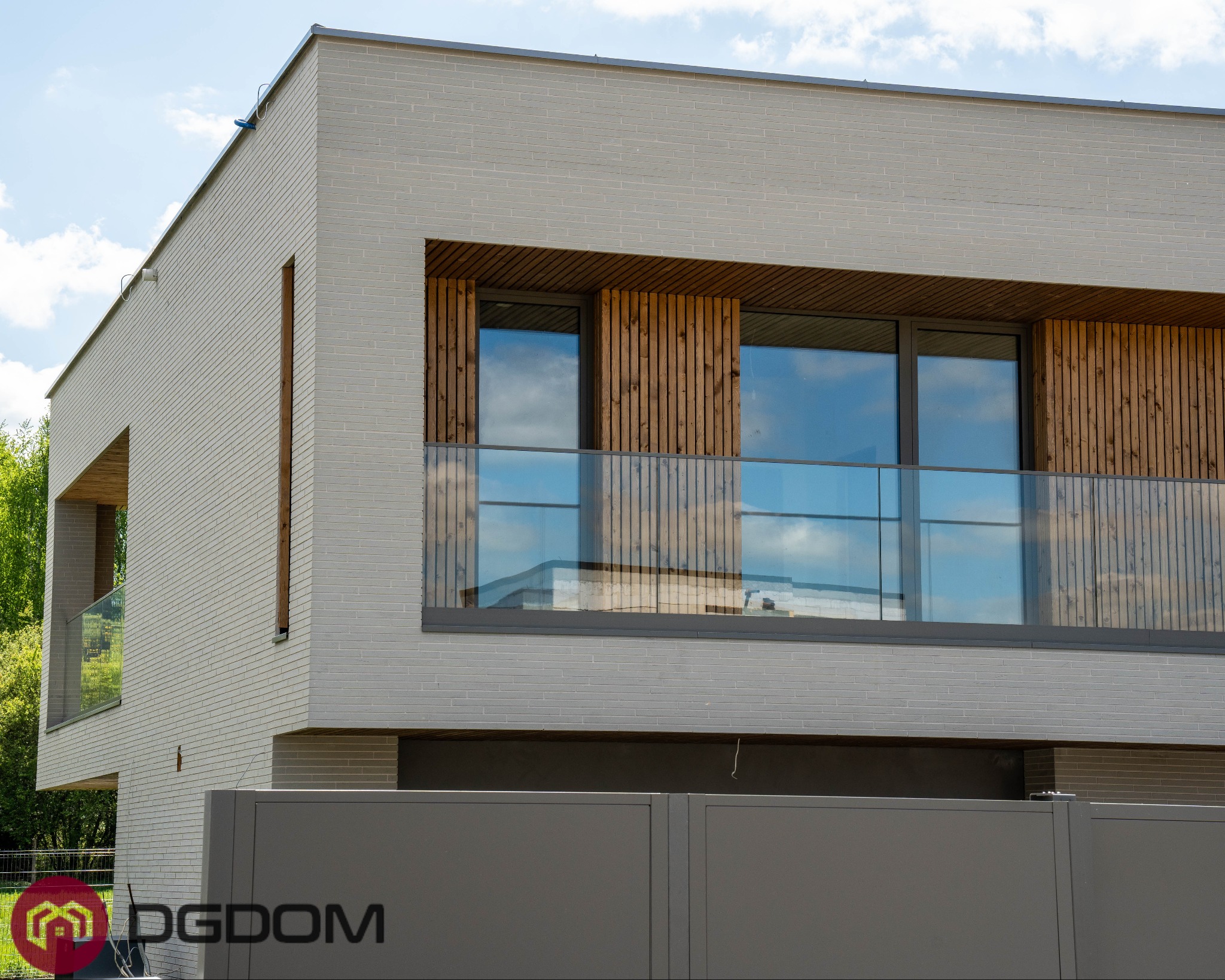 Nowoczesny dom murowany z jasnej cegły, z drewnianymi elementami elewacji i szklaną balustradą balkonu. Widoczny fragment ogrodzenia i zieleń w tle.