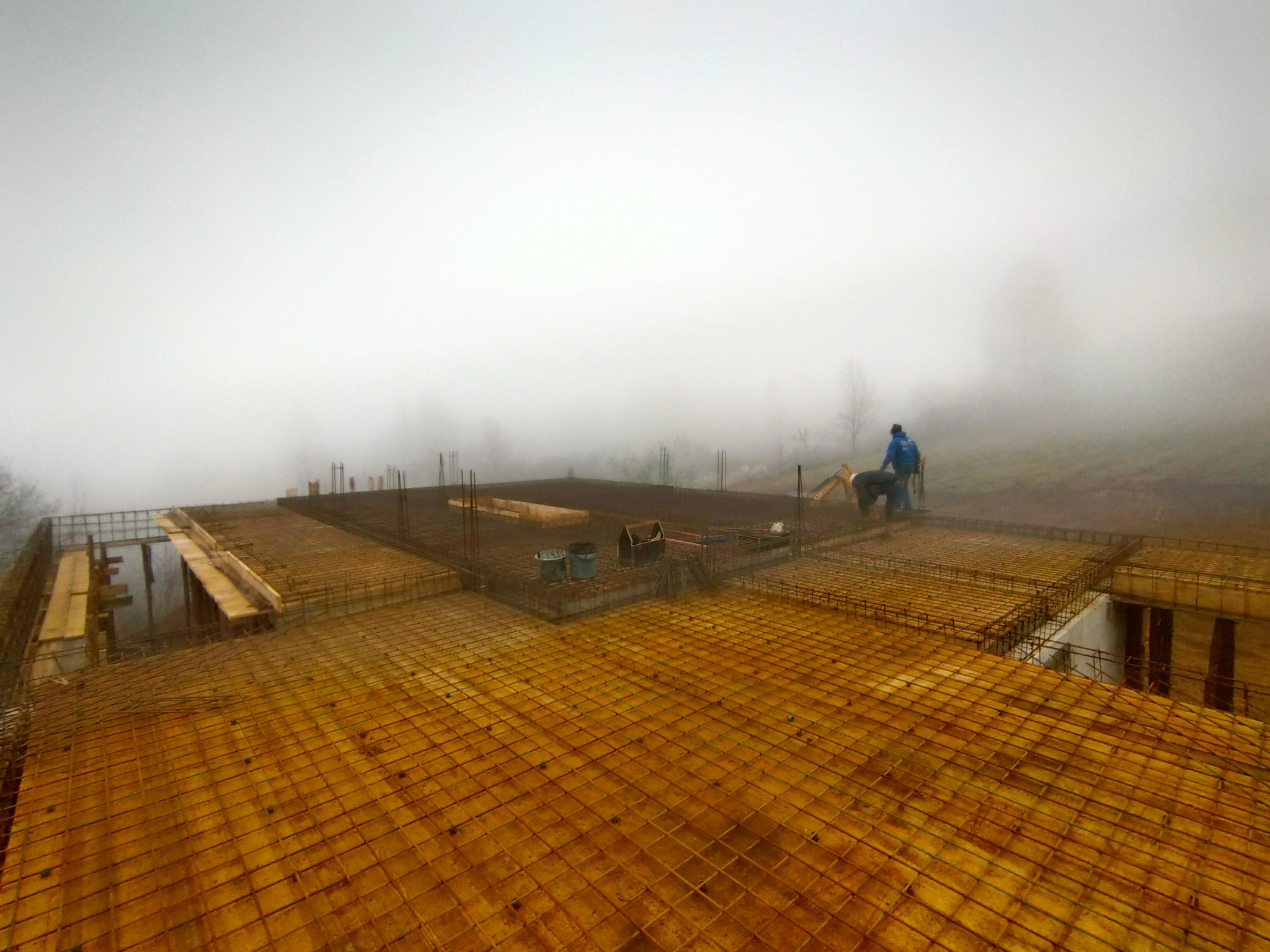 Zbrojenie stropu na budowie domu w mglisty dzień, widoczne siatki zbrojeniowe, betonowe słupy i pracownik w oddali.