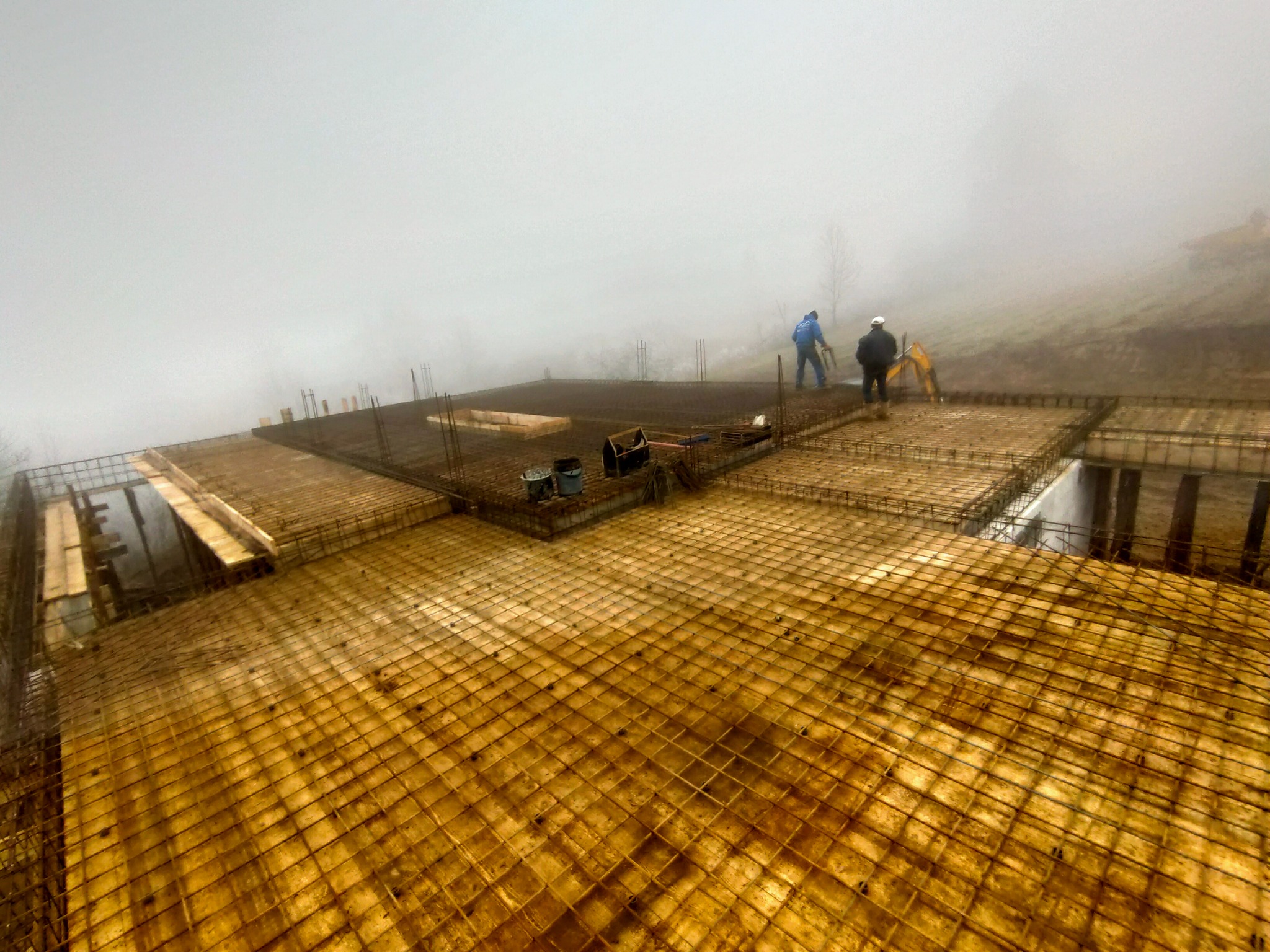 Zalana drewnianym szalunkiem płyta fundamentowa zbrojona stalą, widoczni dwaj pracownicy w oddali oraz koparka w gęstej mgle.