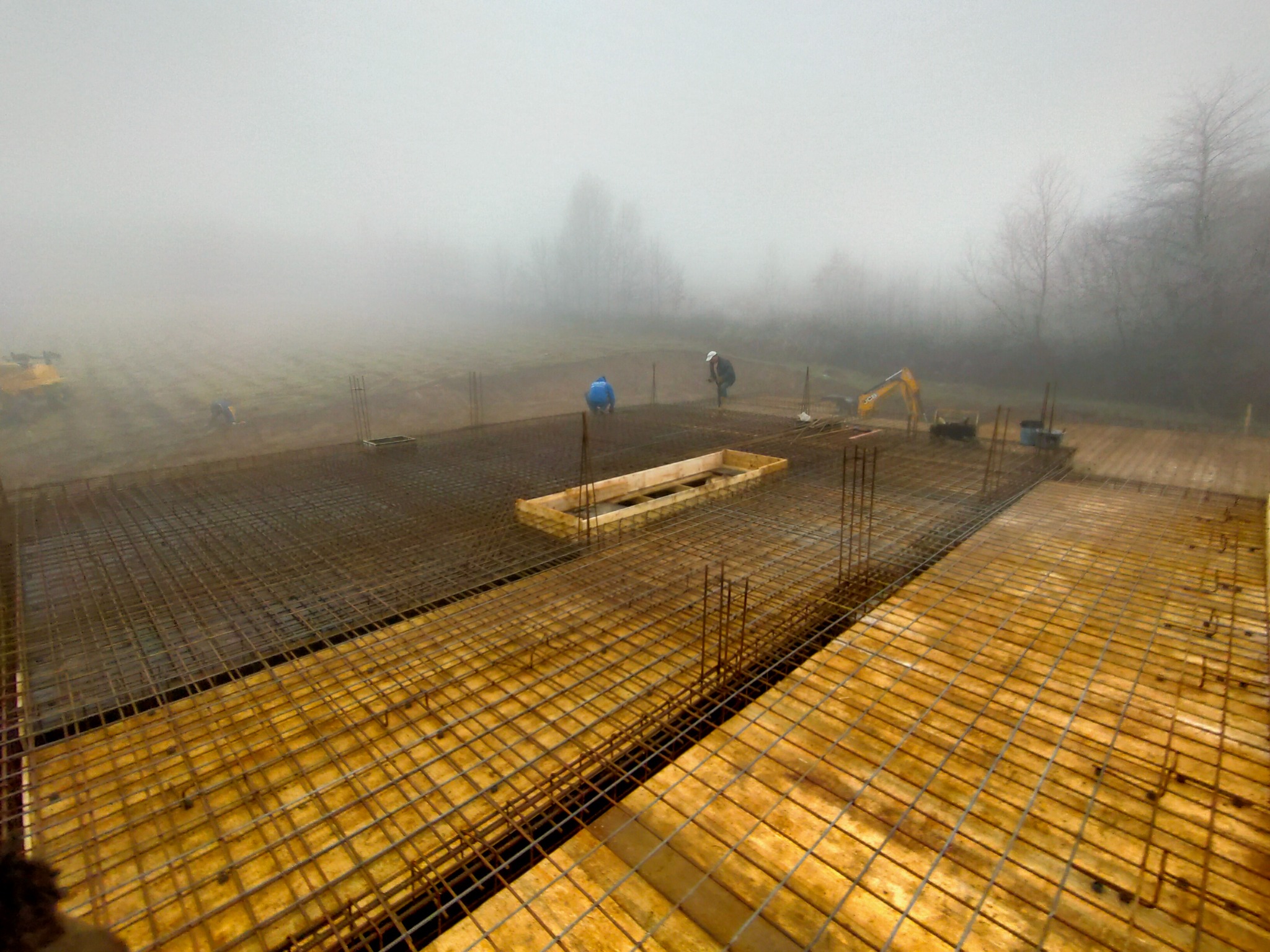 Zbrojenie fundamentów pod dom w gęstej mgle, widoczni robotnicy i koparka w tle.