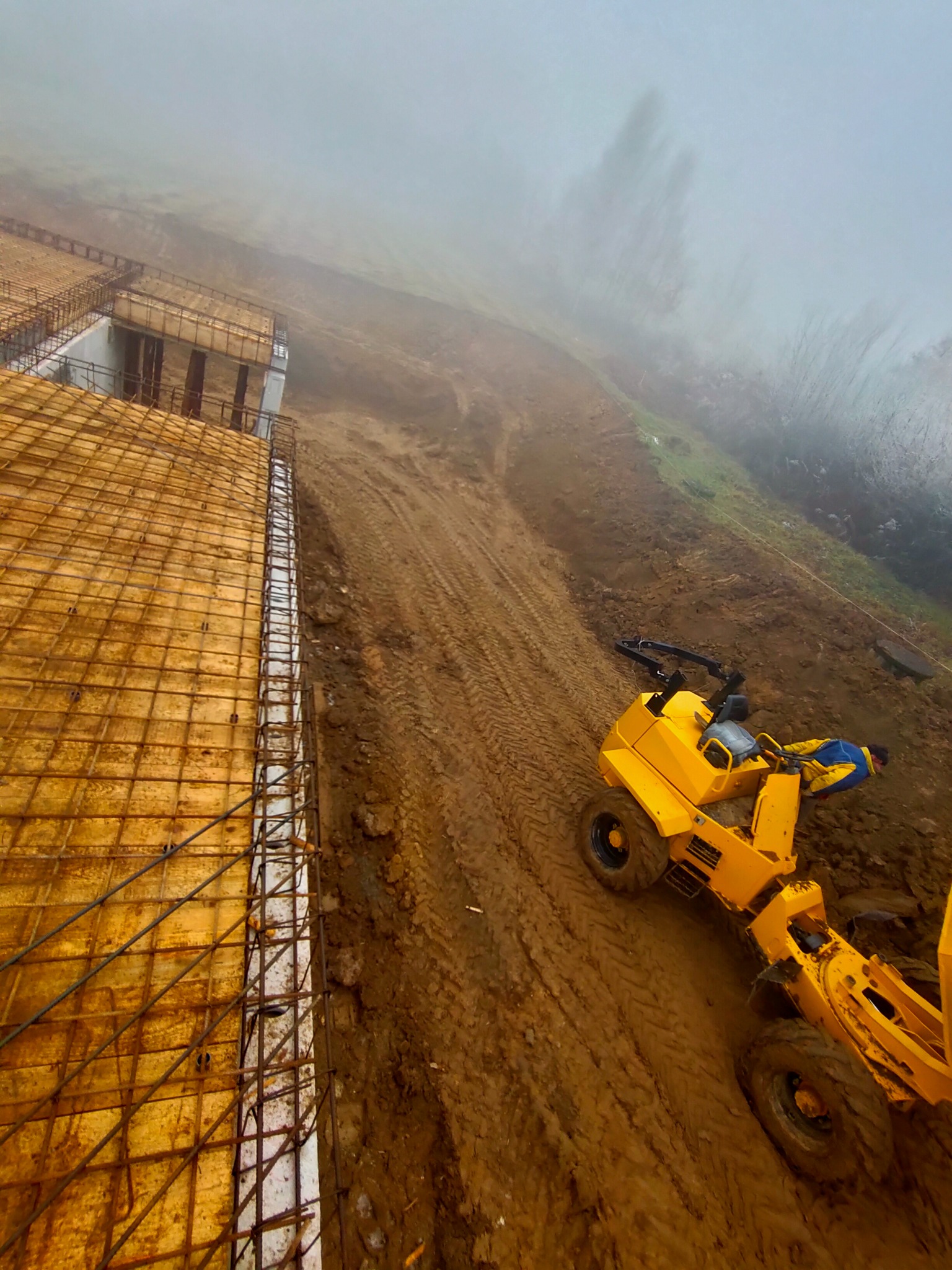 Fundamenty pod budynek w trakcie wylewania, widoczna konstrukcja zbrojeniowa i szalunki, obok żółta maszyna budowlana na błotnistym terenie w mglisty dzień.