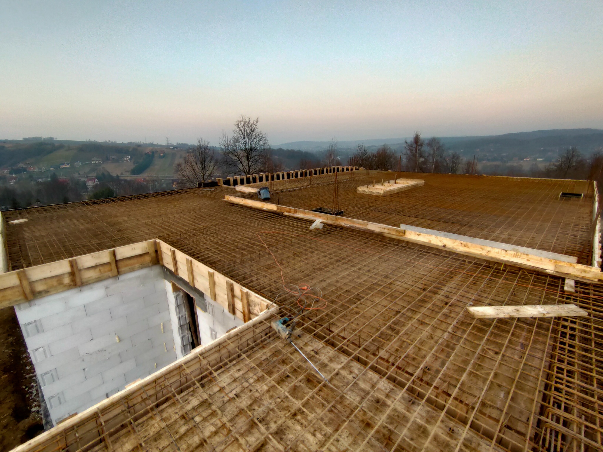 Zbrojenie stropu domu murowanego w trakcie budowy, widok z góry na siatkę z prętów stalowych i szalunki, w tle krajobraz pagórkowaty o zmierzchu.