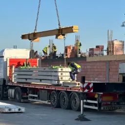 Rozładunek betonowych płyt stropowych z ciężarówki za pomocą dźwigu na placu budowy, pracownicy w kamizelkach odblaskowych nadzorują proces.