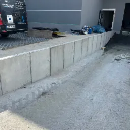 Wzniesiony mur oporowy z prefabrykatów betonowych na placu budowy, widoczny samochód dostawczy firmy z branży budowlanej i materiały budowlane.