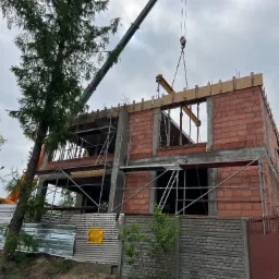 Budowa dwupiętrowego domu z czerwonej cegły, z użyciem dźwigu do podnoszenia belek, widoczne rusztowania i tablica informacyjna na ogrodzeniu.