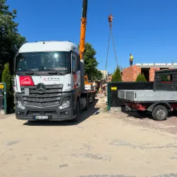 Ciężarówka z dźwigiem firmy Tombud na placu budowy domu z czerwonej cegły, gdzie pracownik w kamizelce stoi na stropie.