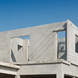 Surowy stan konstrukcji budynku z betonu, widoczne otwory okienne i drzwiowe, wzmocnienia w ścianach, błękitne niebo w tle.