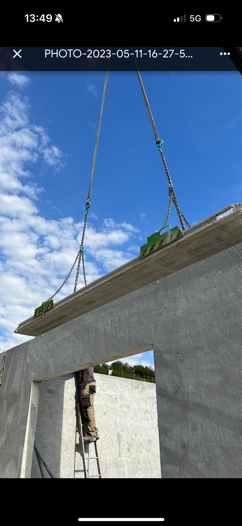 Montaż betonowej płyty stropowej za pomocą łańcuchów i żurawia na budowie domu, widoczny pracownik na drabinie w otworze okiennym, błękitne niebo w tle.