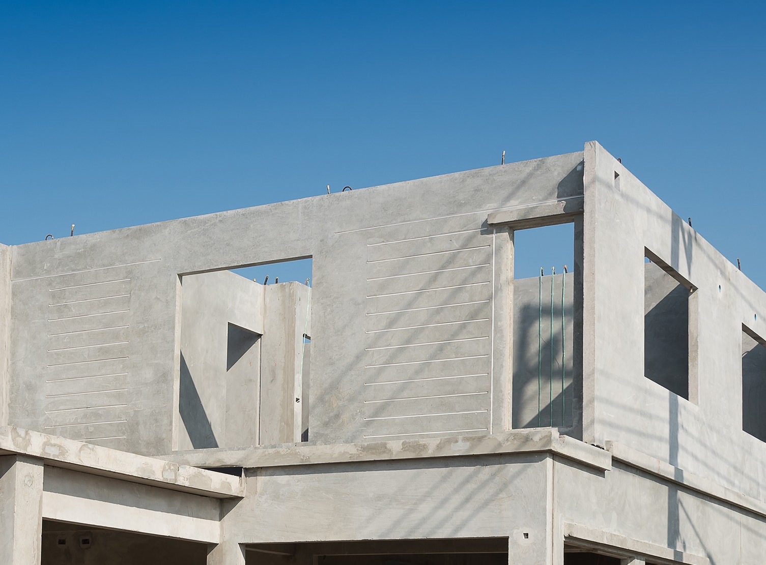 Surowy stan konstrukcji budynku z betonu, widoczne otwory okienne i drzwiowe, wzmocnienia w ścianach, błękitne niebo w tle.