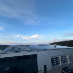 Widok z góry na duży budynek przemysłowy z zainstalowanymi panelami słonecznymi na dachu, błękitne niebo z delikatnymi chmurami w tle, na dole widoczne fragmenty parkingu i zaparkowany czarny...