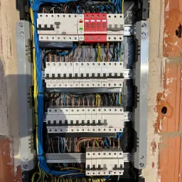 Otwarta skrzynka rozdzielcza z zainstalowanymi bezpiecznikami, przewodami w kolorach niebieskim, żółtym i brązowym, zamontowana w surowej ścianie z cegły.