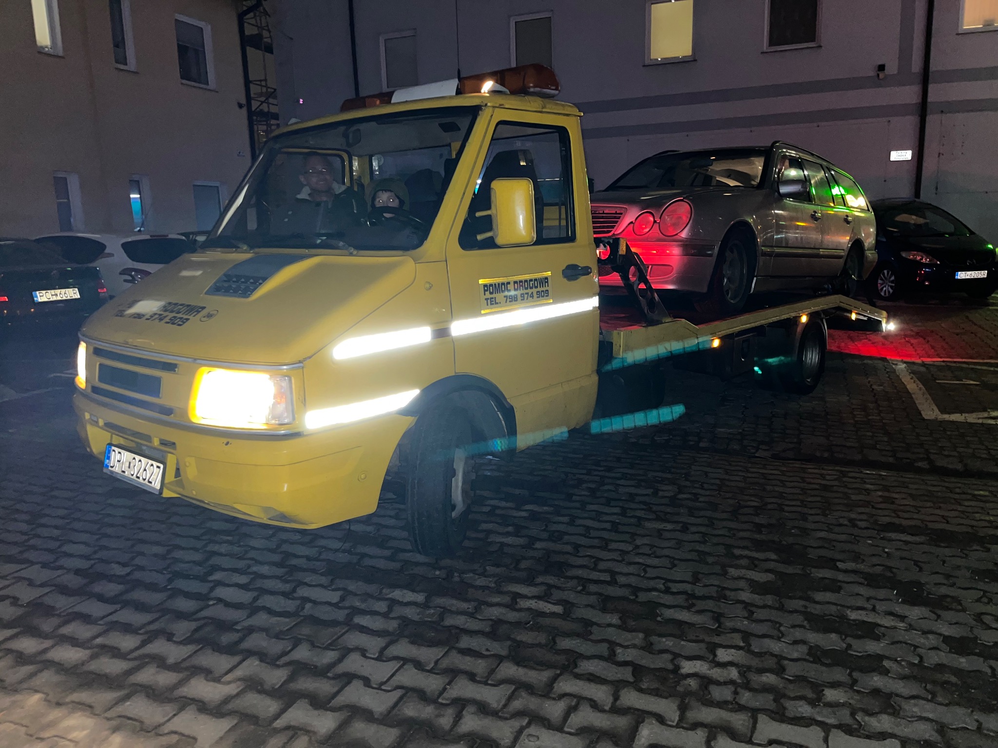 Żółta ciężarówka pomocy drogowej z załadowanym srebrnym kombi Mercedes na platformie. Widoczni kierowca i pasażer w kabinie, w tle budynek i zaparkowane samochody, nocne oświetlenie.