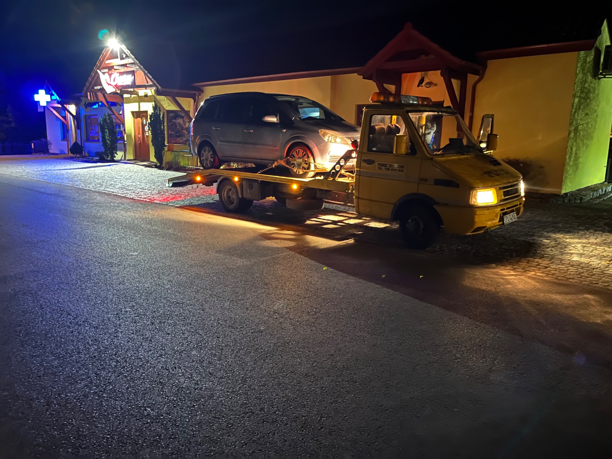 Żółta laweta z załadowanym srebrnym minivanem na tle oświetlonego budynku z niebieskim krzyżem aptecznym, wykonana nocą.