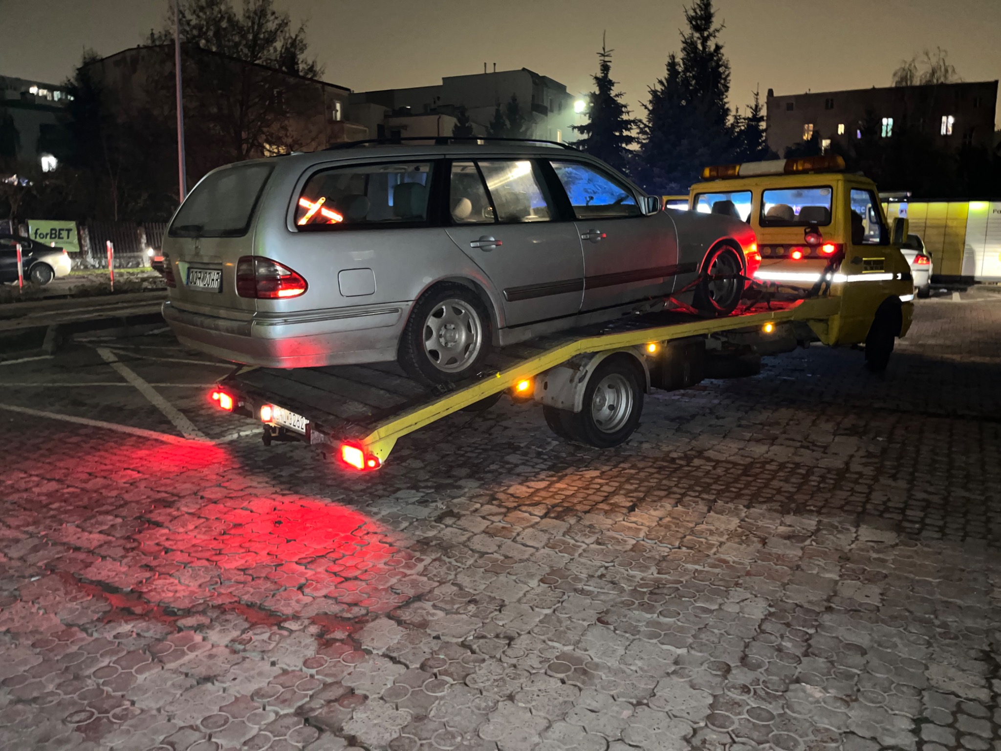 Srebrne kombi Mercedes na żółtej lawecie, z czerwoną taśmą w kształcie krzyża w tylnej szybie, oświetlone czerwonymi światłami lawety na tle nocnego parkingu miejskiego.
