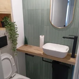 Nowoczesna, minimalistyczna łazienka z zielonymi płytkami ułożonymi pionowo, drewnianym blatem z białą umywalką nablatową i czarną armaturą, lustro w złotej ramie, toaleta z otwartą klapą...