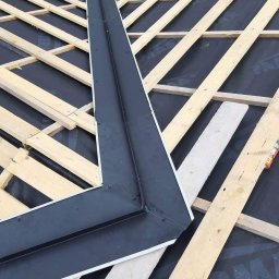 Usługi Dekarskie Blacharskie Naprawa Dachow - Wiązka drewnianych łat na czarnej membranie dachowej z zamontowanym, czarnym, kątowym elementem blacharskim, widoczne fragmenty obuwia dekarza.