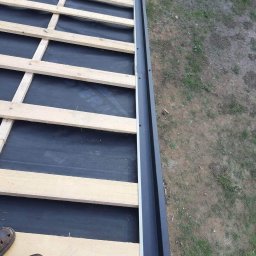 Usługi Dekarskie Blacharskie Naprawa Dachow - Wiązar dachowy z czarną membraną i obróbką blacharską krawędzi dachu, widok z góry z butami dekarza w lewym dolnym rogu.