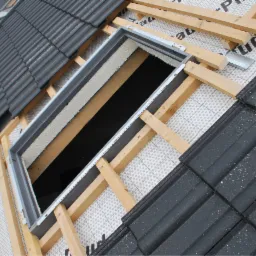 Montaż okna dachowego w trakcie układania dachówki ceramicznej; widoczne drewniane łaty, membrana dachowa z nadrukiem oraz metalowa rama okna.