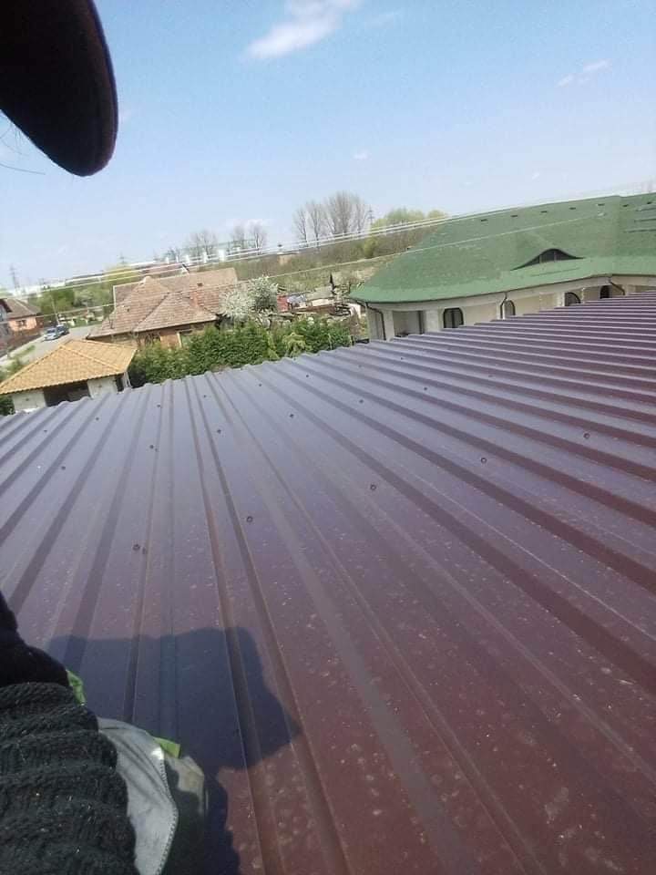 Nowe, brązowe, blaszane pokrycie dachu z widocznymi nitami mocującymi, na tle zabudowy jednorodzinnej i błękitnego nieba.